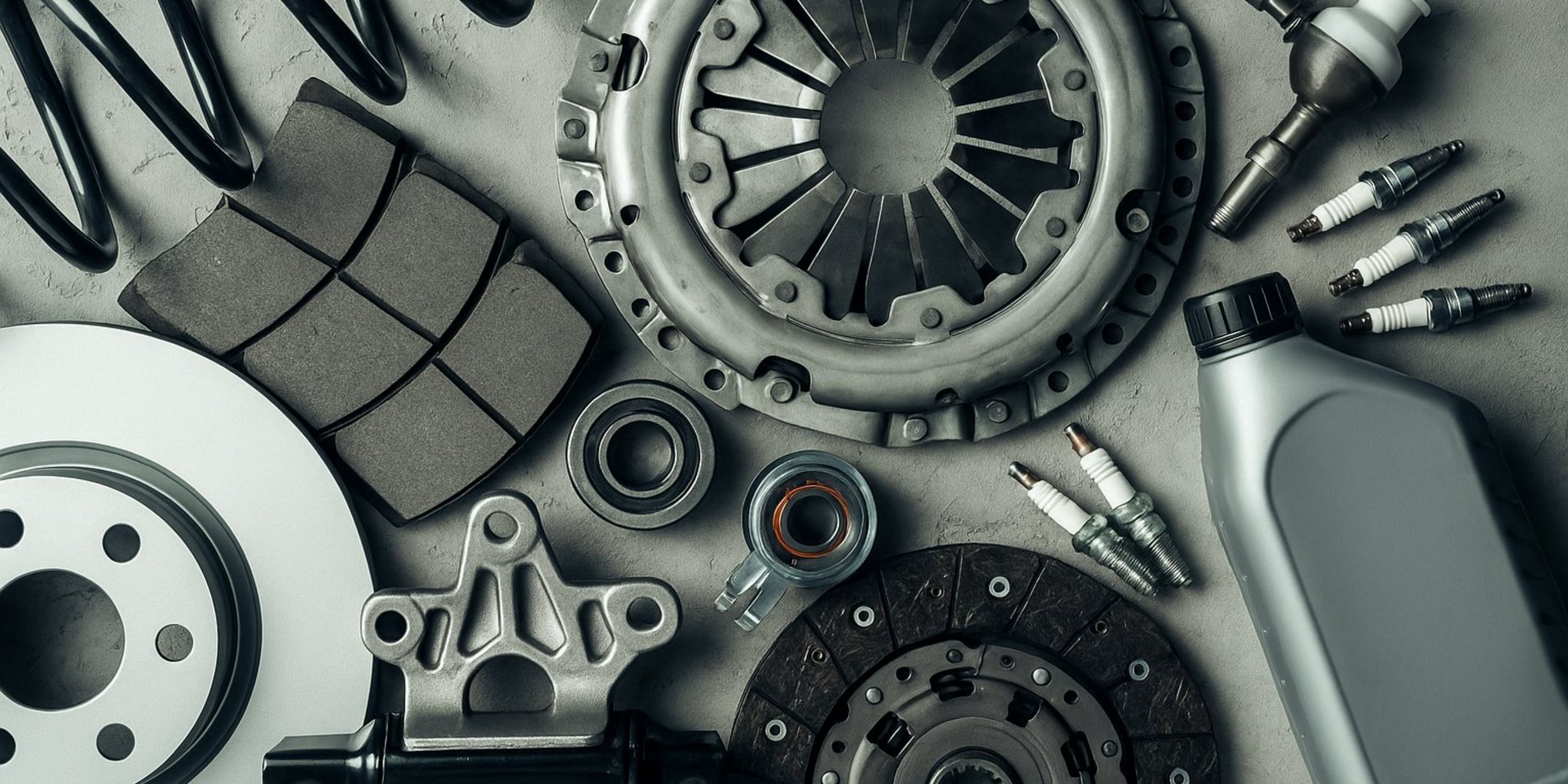 Verschiedene Autoersatzteile auf grauem Untergrund arrangiert, darunter eine Kupplungsscheibe, ein Drucklager, Zündkerzen, ein Stoßdämpfer, Bremsbeläge, ein Bremsscheibe, ein Kugellager, eine Flasche Motoröl und eine Schraubenfeder – flach aus der Vogelperspektive fotografiert.