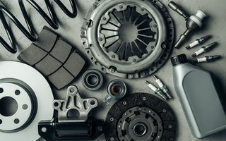 Verschiedene Autoersatzteile auf grauem Untergrund arrangiert, darunter eine Kupplungsscheibe, ein Drucklager, Zündkerzen, ein Stoßdämpfer, Bremsbeläge, ein Bremsscheibe, ein Kugellager, eine Flasche Motoröl und eine Schraubenfeder – flach aus der Vogelperspektive fotografiert.