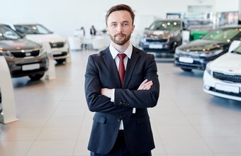 Ein Mann im Anzug steht mit verschränkten Armen in einem Autohaus vor mehreren Fahrzeugen.