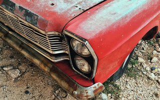 Detailaufnahme eines alten, verrosteten roten Autos mit verblasstem Lack und beschädigter Stoßstange.